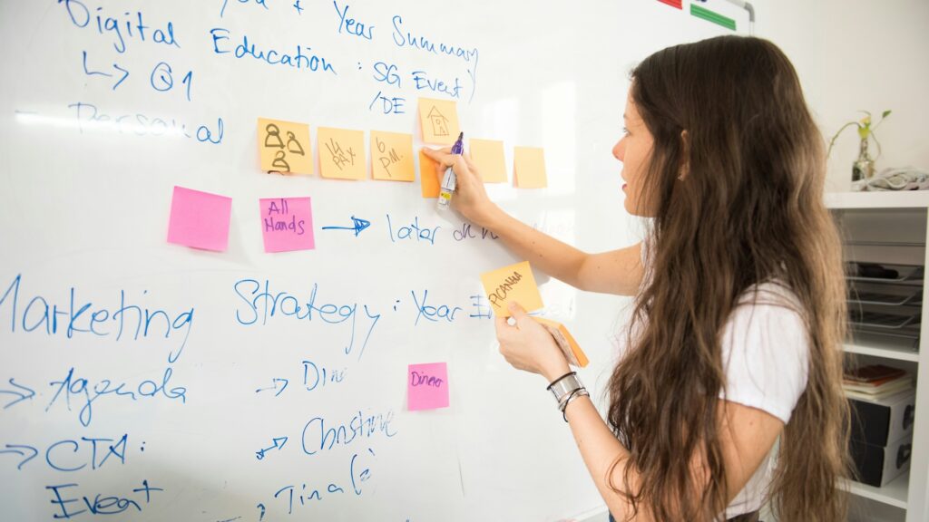 En kvinna står framför en whiteboard och sätter upp postits. Det står ord som digital och marketing, och kvinnan ser allvarlig eller fundersam ut.