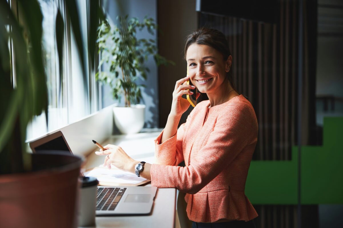 Bild på en glad CMO eller marknadschef som tittar rakt in i kameran. Hon pratar i telefon klädd i en aprikos kavaj och ser avslappnad ut.